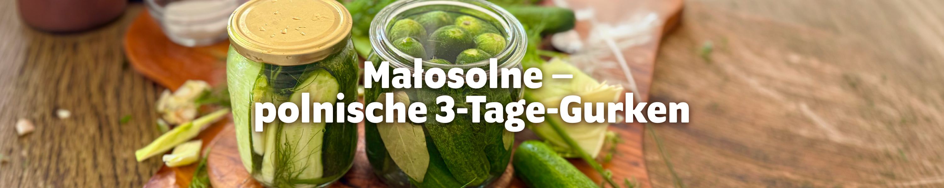 Zwei Einmachgläser mit frisch fermentierten Małosolne-Gurken auf einem Holzbrett, umgeben von Dill, Lorbeerblättern und frischem Knoblauch – Header-Bild zum Rezept „Małosolne – polnische 3-Tage-Gurken"